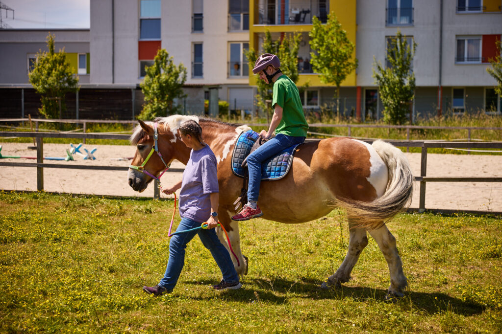 Ein Schüler mit Beeinträchtigungen reitet auf einem Pferd, das von der verantwortlichen Lehrkraft für das heiltherapeutische Reiten geführt wird.